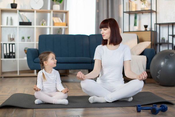 Happy mother with little daughter meditating, sitting in easy seat pose on mat at home, smiling caring young mom with adorable girl child practicing yoga together, healthy lifestyle concept. - Zdjęcie, obraz