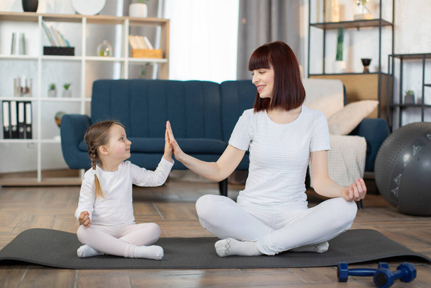 Family, mother and child daughter are engaged in fitness, yoga, exercise at home. Smiling young red hair woman looking at small kid girl sitting on the carpet floor in lotus pose and giving high five. - Foto, Bild