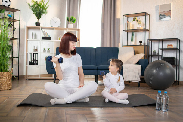 Sport, workout concept. Mom and Little Girl doing dumbbell exercises on a yoga mat in the living room at home. New normal concept. Healthy lifestyle. Relaxing at home. - Foto, afbeelding