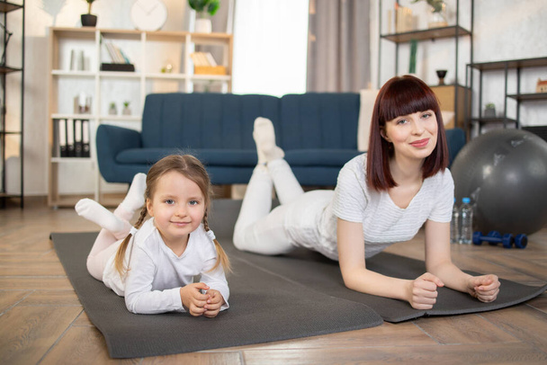 A young caucasian mother and her little daughter lying on a mat on the floor during fitness classes in the living room at home and smiling at the camera. Healthy Lifestyle - Foto, afbeelding