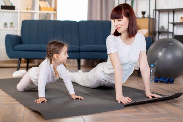 A young caucasian mom with her little daughter doing fitness, stretching, push up exercises on the floor mat in the living room. Sports at home and female beauty. - Foto, imagen