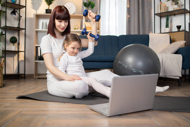 Young beautiful mother sitting on a gray mat on floor with a little charming daughter in white sportswear, doing exercises with dumbbells while looking online fitness course on a laptop. - Фото, зображення