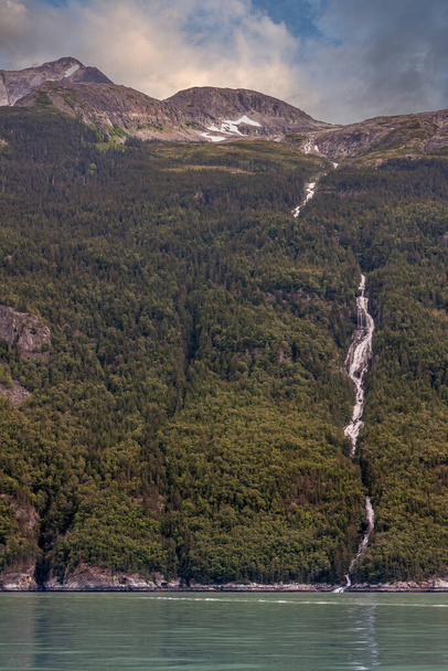 Skagway, Alaska, USA - July 20, 2011: Taiya Inlet above Chilkoot Inlet. Wide view on waterfall from top of mountain till reaching green ocean water under blue cloudscape. - Photo, Image