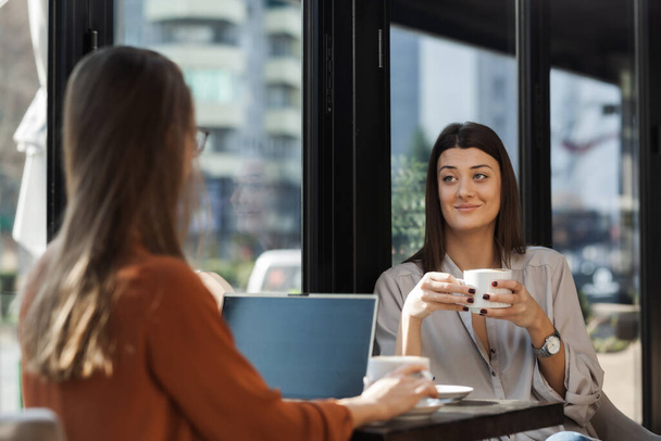 Two young business women in a cafe having one on one meeting. Friends after work talking gossiping and having coffee at a window table on a sunny day. - Foto, Imagem