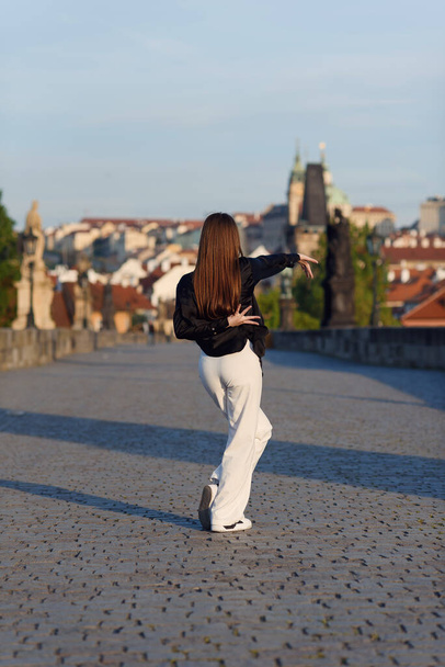 Young attractive woman in casual stylish clothes dancing on the stone bridge of old town in a Prague. Active lifestyle and dancing concept. - Foto, afbeelding