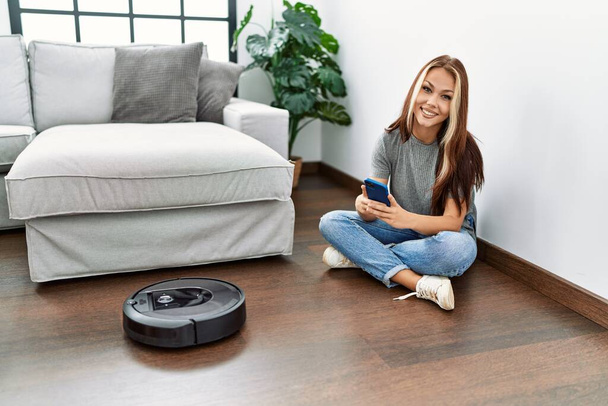 Young caucasian girl controlling robot vacuum cleaner with smartphone sitting on the floor at home. - Photo, Image