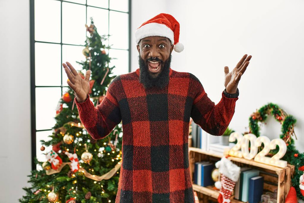 Αφροαμερικάνος που φοράει καπέλο Σάντα Κλάους στέκεται δίπλα στο χριστουγεννιάτικο δέντρο γιορτάζοντας την τρέλα και έκπληκτος για την επιτυχία με τα χέρια σηκωμένα και τα ανοιχτά μάτια ουρλιάζοντας ενθουσιασμένος. έννοια νικητή  - Φωτογραφία, εικόνα