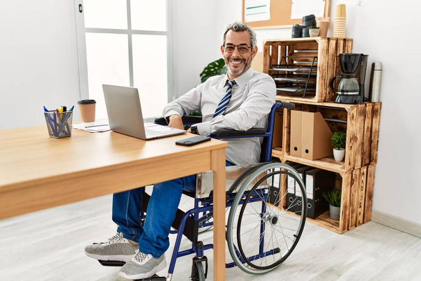 Middle age hispanic man working at the office sitting on wheelchair looking away to side with smile on face, natural expression. laughing confident.  - Photo, Image