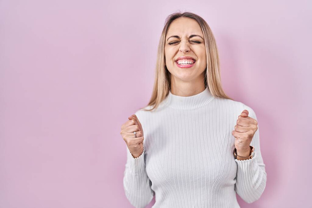 Junge blonde Frau in weißem Pullover vor rosa Hintergrund begeistert für den Erfolg mit erhobenen Armen und geschlossenen Augen feiern den Sieg lächelnd. Siegerkonzept.  - Foto, Bild