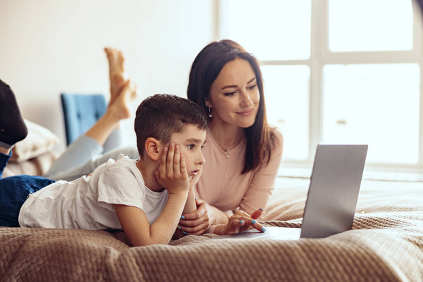 Attractive Caucasian mother and small teen son lying on bed and looking at laptop screen. Portrait. - Foto, imagen