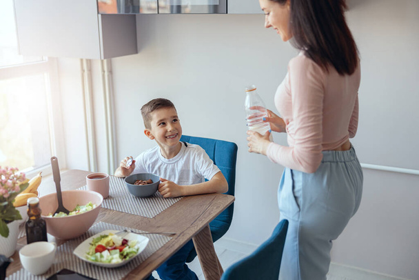 Küçük beyaz çocuk mutfak masasında mısır gevreğiyle oturuyor, annesi sütlü. Kahvaltı.. - Fotoğraf, Görsel