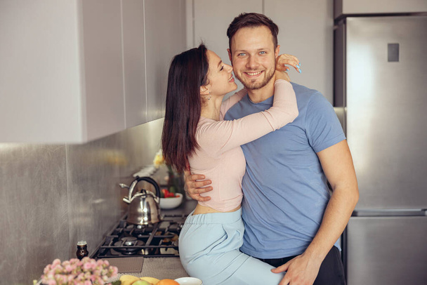 Portrait of happy Caucasian young handsome man in kitchen in hugs of beloved woman. Couple in love. - Photo, Image