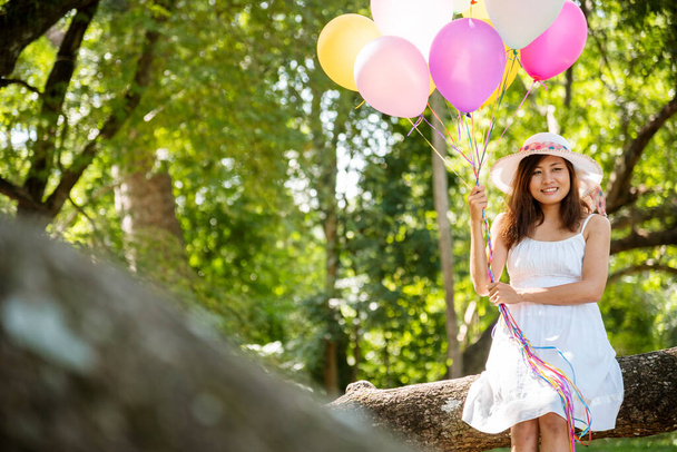Fröhliche Schönheitskönigin mit Luftballons entspannt unter einem großen Baum im grünen Park sitzend. Frau Hände, die lebendige Luftballons halten, spielen auf Geburtstagsparty fröhliche Zeit Sommer bei Sonnenschein im Freien - Foto, Bild