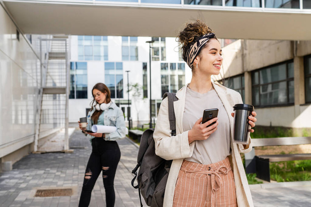Deux étudiantes marchent devant le bâtiment de l'université dans l'espace de copie de jour femme caucasienne utilisant un téléphone portable marchant devant le bâtiment moderne portant des brunes occasionnelles de vraies personnes - Photo, image