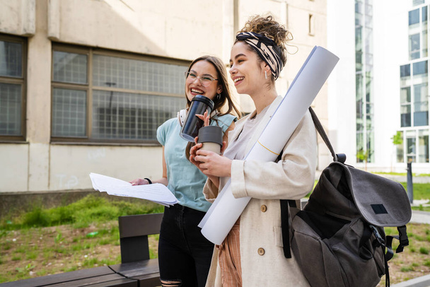 Дві жінки-студенти, які ходять разом перед будівлею університету в літній день, тримають нотатки і говорять про іспит або урок реальні люди кавказькі колеги копіюють простір
 - Фото, зображення