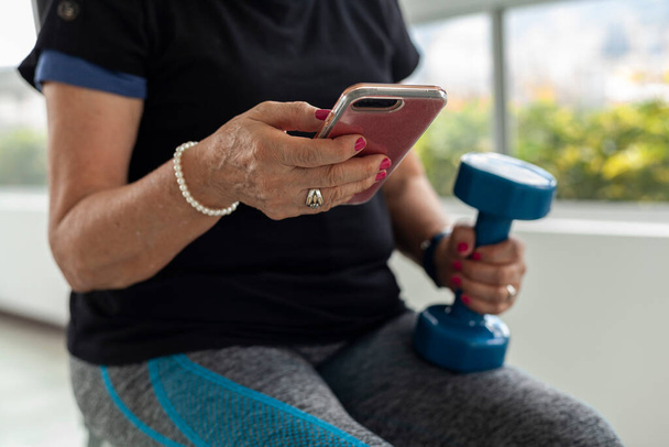 Older Latina woman works out at gym with dumbbells using cell phone workout videos - Foto, afbeelding