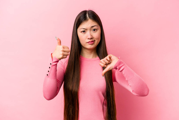 Fiatal kínai nő elszigetelt rózsaszín háttér mutatja hüvelykujj fel és hüvelykujj le, nehéz választani koncepció - Fotó, kép