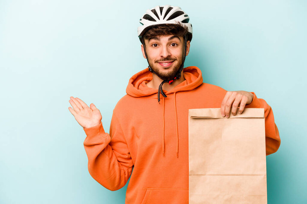 Nuori latino mies yllään kypärä pyörä tilalla ottaa pois ruokaa eristetty sinisellä pohjalla osoittaa kopio tilaa kämmenellä ja tilalla toinen käsi vyötäröllä. - Valokuva, kuva