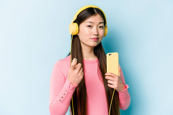 Mladá asijská žena poslouchá hudbu izolované na modrém pozadí ukazuje prstem na vás, jako by pozvání přijít blíž. - Fotografie, Obrázek