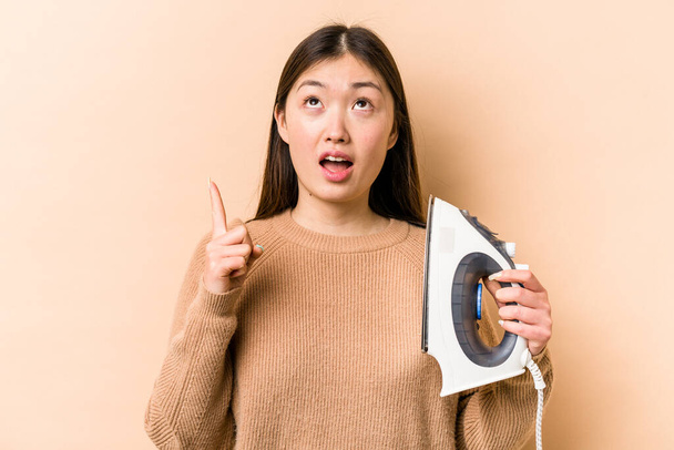 Young asian woman holding iron isolated on beige background pointing upside with opened mouth. - Fotografie, Obrázek