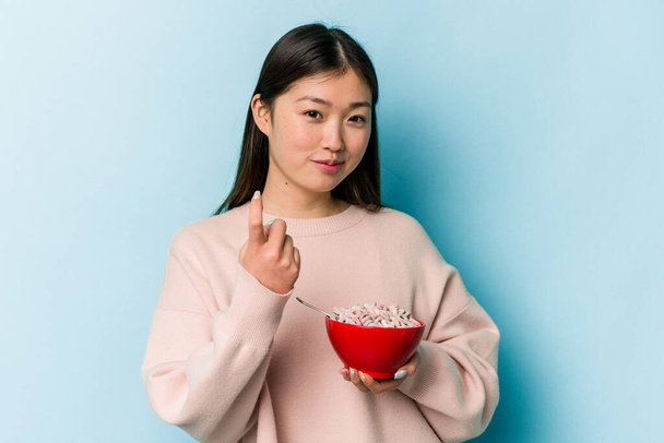 Giovane donna asiatica in possesso di una ciotola di cereali isolato su sfondo blu che punta con il dito a voi come se invitante avvicinarsi. - Foto, immagini