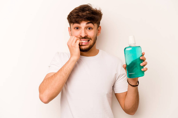 Young hispanic man holding mouthwash isolated on white background biting fingernails, nervous and very anxious. - Photo, image