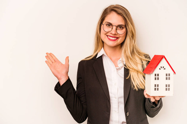 Young business caucasian woman holding a toy house isolated on white background showing a copy space on a palm and holding another hand on waist. - Photo, Image