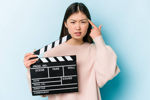 jonge aziatische vrouw houden clapperboard geïsoleerd op blauwe achtergrond tonen een teleurstelling gebaar met wijsvinger. - Foto, afbeelding
