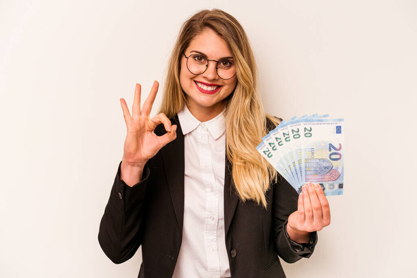 Young business caucasian woman holding a banknotes isolated on white background cheerful and confident showing ok gesture. - Photo, Image