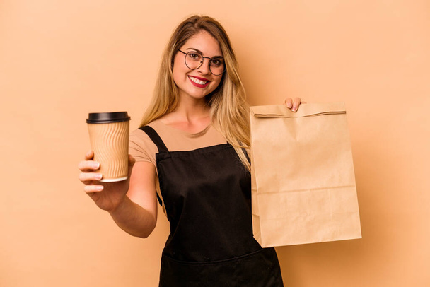 Restaurant waiter caucasian woman holding a take away coffee and bag isolated on beige background - Φωτογραφία, εικόνα