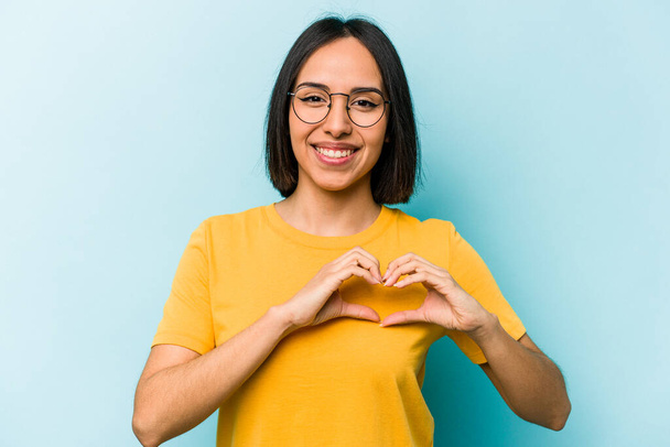 Молодая латиноамериканка на синем фоне улыбается и показывает форму сердца руками. - Фото, изображение