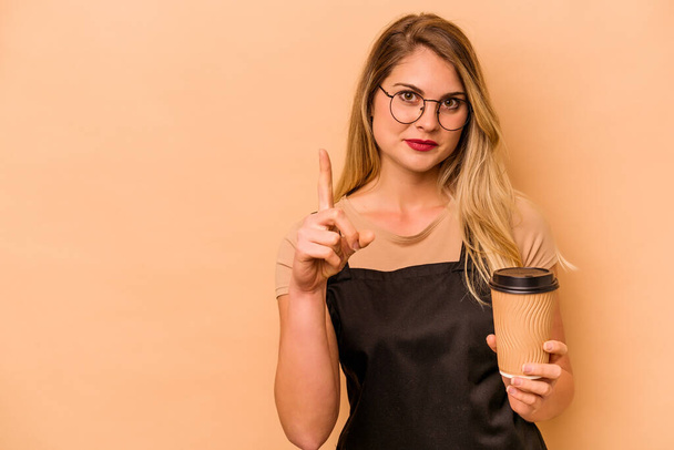 Ristorante cameriere donna caucasica in possesso di un caffè da asporto isolato su sfondo beige mostrando il numero uno con il dito. - Foto, immagini