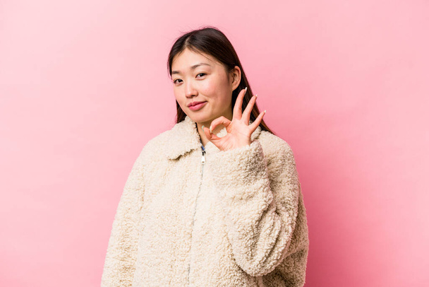 Giovane donna cinese isolata su sfondo rosa ammicca un occhio e tiene un gesto corretto con la mano. - Foto, immagini
