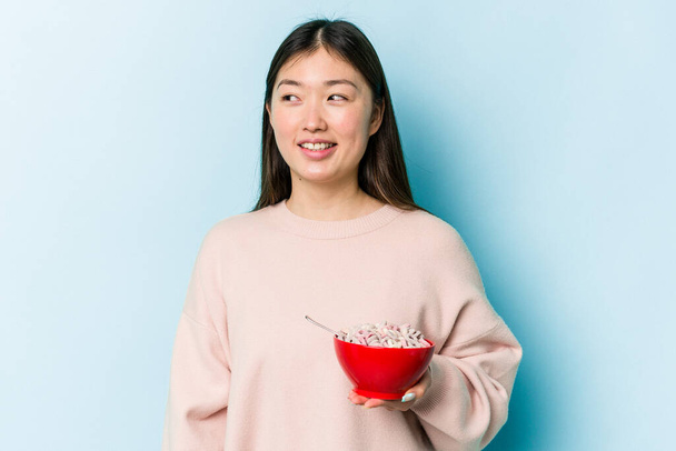 Junge asiatische Frau hält eine Schale mit Getreide isoliert auf blauem Hintergrund und blickt lächelnd, fröhlich und angenehm zur Seite. - Foto, Bild