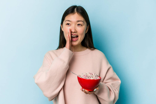 Joven mujer asiática sosteniendo un tazón de cereales aislado sobre fondo azul gritando y sosteniendo la palma cerca de la boca abierta. - Foto, Imagen