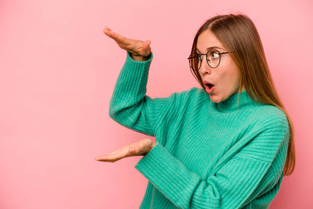 Young caucasian woman isolated on pink background shocked and amazed holding a copy space between hands. - Photo, Image