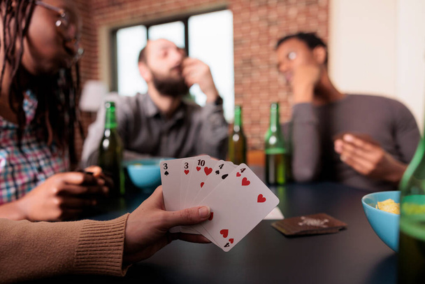 Κοντινό πλάνο της γυναίκας χέρι κρατώντας παιχνίδια καρτών, ενώ απολαμβάνοντας διασκεδαστική δραστηριότητα αναψυχής με τους καλύτερους φίλους. Πρόσωπο που παίζει χαρτιά παιχνίδια με ανθρώπους που έχουν σνακ και ποτά, ενώ κάθεται στο σπίτι. - Φωτογραφία, εικόνα
