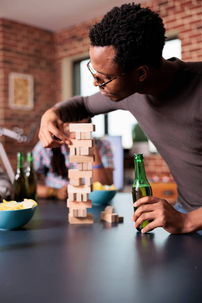 Focused african american man carefully removing wood block from wooden tower structure. Concentrated confident person sitting at table in living room while playing society games with friends. - Фото, зображення