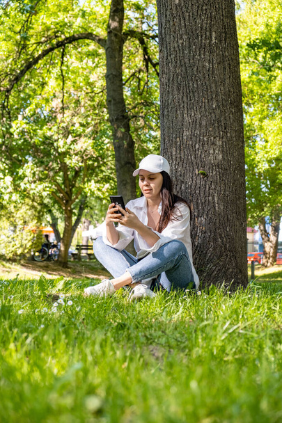 woman sitting under the tree at city park surfing internet on the phone copy space - Photo, Image