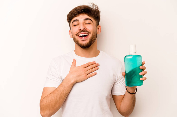 Jonge Spaanse man houdt mondwater geïsoleerd op een witte achtergrond lacht luid hand in hand op de borst. - Foto, afbeelding