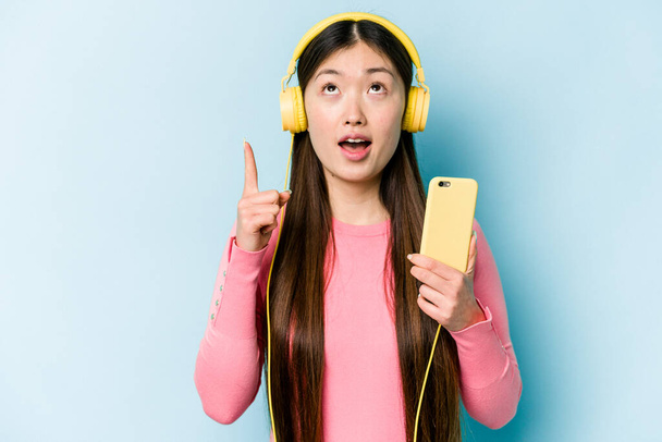 Jovem mulher asiática ouvindo música isolada no fundo azul apontando de cabeça com a boca aberta. - Foto, Imagem