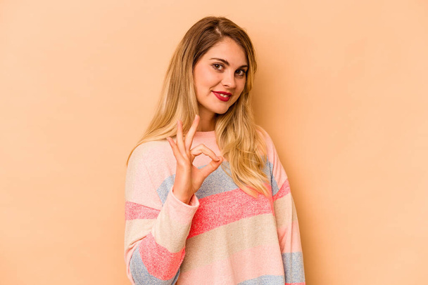 Young caucasian woman isolated on beige background winks an eye and holds an okay gesture with hand. - Photo, Image