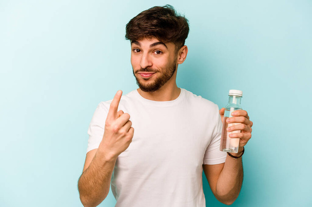 Jovem hispânico segurando uma garrafa de água isolada no fundo branco apontando com o dedo para você como se convidando chegar mais perto. - Foto, Imagem