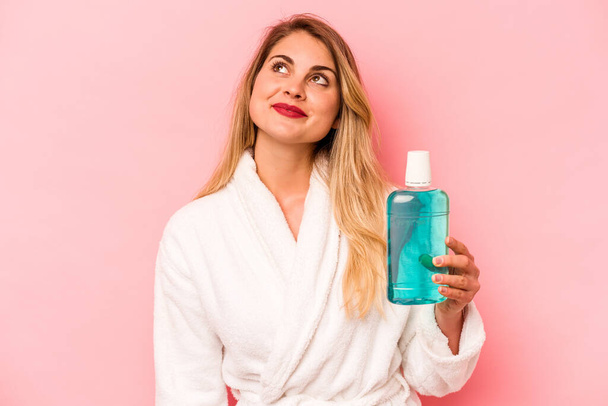 Young caucasian woman holding mouthwash wearing bathrobe isolated on pink background dreaming of achieving goals and purposes - Photo, image