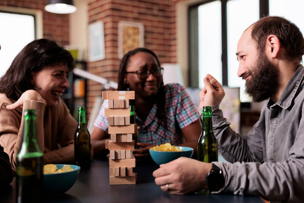 Lachen diverse mensen zitten aan tafel terwijl praten en genieten van de samenleving games samen. Multi-etnische mensen zitten thuis in de woonkamer tijdens het bespreken en spelen met houten blokken. - Foto, afbeelding