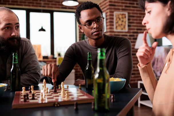 Στραγγαλισμένοι πολυεθνικοί άνθρωποι κάθονται στο τραπέζι παίζοντας σκάκι μαζί. Πολυφυλετικές επικεντρώθηκε φίλους κάθεται στο σπίτι στο σαλόνι, ενώ απολαμβάνοντας επιτραπέζιο παιχνίδι στρατηγικής και σνακ. - Φωτογραφία, εικόνα