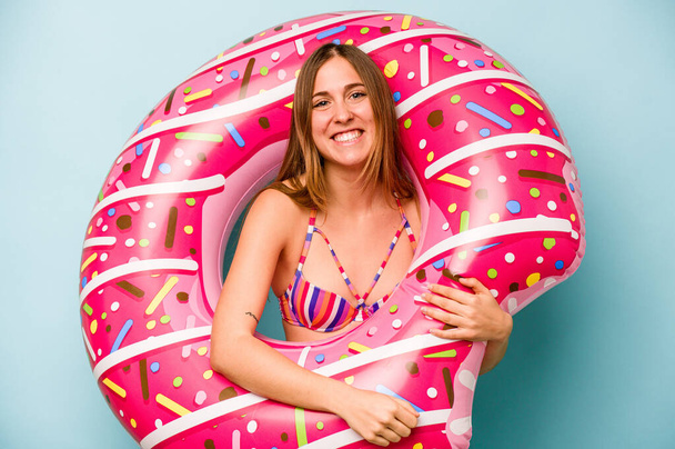 Young caucasian woman holding air mattress isolated on blue background laughing and having fun. - Photo, Image