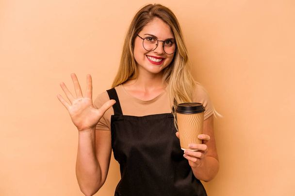 Restoran garsonu Kafkasyalı kadın bej arka planda izole edilmiş kahve tutuyor. Beş numarayı parmaklarıyla gülümsüyor.. - Fotoğraf, Görsel