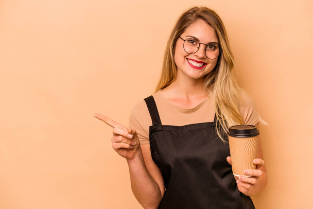 Serveur restaurant femme caucasienne tenant un café à emporter isolé sur fond beige souriant et pointant de côté, montrant quelque chose à vide. - Photo, image