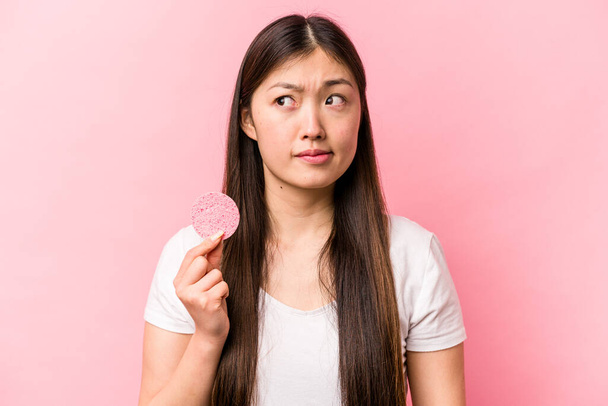 Молодая азиатская женщина с губкой на лице изолированы на розовом фоне путают, чувствует себя сомнительным и неуверенным. - Фото, изображение